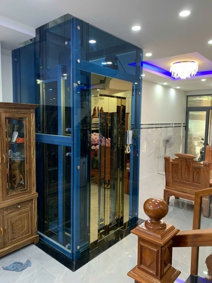 Thang máy khung thép vách kính xanh cửa inox vàng gương khung bao bản rộng